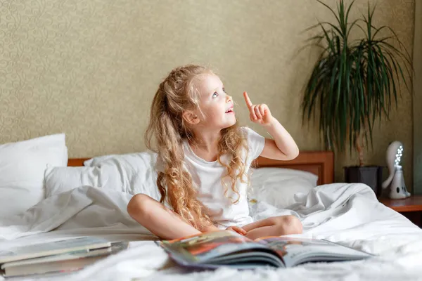 宿題をしている間に朝のベッドで本を読んでいる間に指で指差す賢い美しい小さな子供の女の子は家のインテリアの寝室で想像する勉強を読みます。ブロンド女には賢い考えがある. — ストック写真