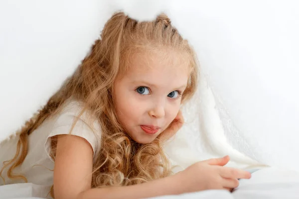 Retrato de bela menina sorridente mostrando língua escondida sob cobertor branco. Brincalhão encantador encaracolado criança loira joga esconder e procurar sob cobertor em casa. — Fotografia de Stock