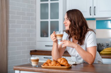 Genç ve güzel bir kadın Cozy 'nin evindeki mutfakta kruvasanla kahve kapuçino içiyor. Gülümseyen 30 'lu yaşlardaki beyaz kadın sabah rutininde kahvaltı yapıyor. Boşluğu kopyala
