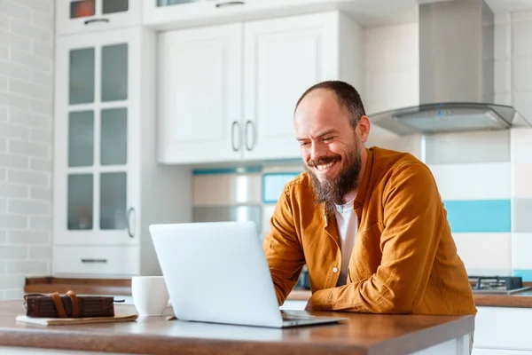 Mladý muž pracující s laptopem v kuchyni v interiéru. Usmívající se Freelancer pracuje vzdáleně z domova. Šťastný majitel dělá papírování s notebookem uvnitř. Plešatý běloch 30s — Stock fotografie