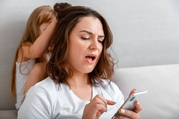 Ibu muda dengan kecanduan internet menggunakan smartphone sementara putri stres yang tidak bahagia menarik rambutnya perkelahian nakal di rumah. Pertengkaran antara anak perempuan dan ibu karena kurangnya perhatian Stok Gambar
