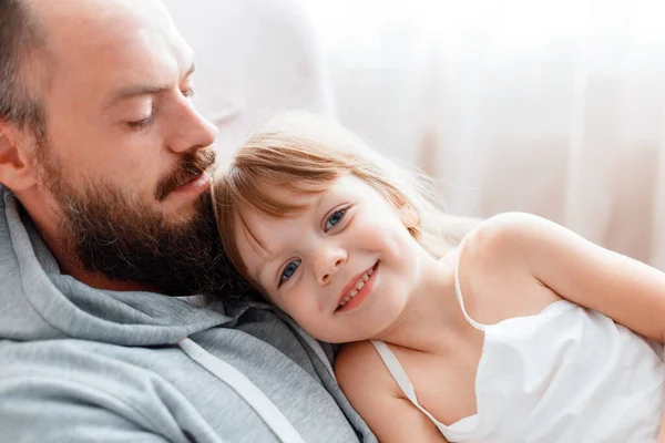 Miúda sorridente a abraçar o papá, mente no peito do pai. Pai carinhoso caucasiano barbudo homem dos anos 30 abraça sua filhinha em casa interior. Feliz paternidade. Imagem De Stock