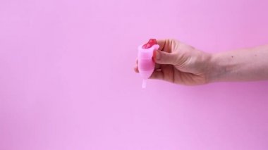 Kadın eli, menstrüal fincanın pembe arka planda kırmızı kurdeleyle gösterildiğini gösteriyor. Adet bardağını tutan kadın. Kadın mahrem hijyen dönemi sıfır atık ürünü. Kadın sağlığı kavramı.