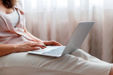 Calm woman use laptop at cozy home interior. Close up female hands typing on keyboard, working using laptop or do online shopping at home on pink chair near window.