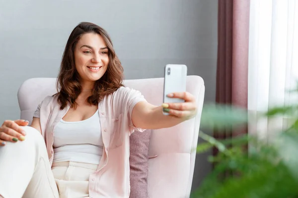 若い優しい笑顔の白人30代の女性が窓の近くの携帯電話を使って自撮り写真を撮っています。長い髪のブルネットはスマートフォンのホームインテリアで自分自身を撮影しています. — ストック写真