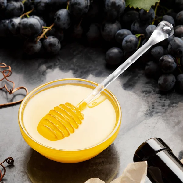Honey in glass jar with honey spoon on dark stone board with wine cheese corkscrew in Black juicy grapes frame. Delicious yellow bee honey in snack plate on black table. Square photo — Stock Photo, Image