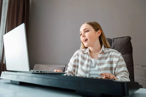 Woman learns music singing vocals playing piano online using laptop at home interior. Teenager girl sings song and plays piano synthesizer during video call, online lesson with teacher. Лицензионные Стоковые Фото