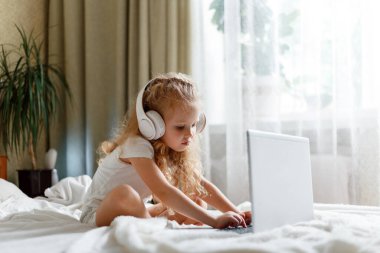 Anaokulundaki kulaklıklı kız, anaokulunda uzaktan eğitim için dizüstü bilgisayar kullanıyor. Evde yatağında ödev yaparken dizüstü bilgisayar kullanan akıllı sakin kız çocuğu. çocuklar için e-öğrenme