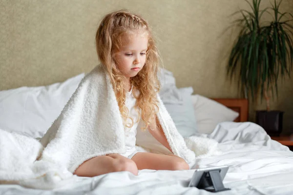 家庭でテレビアニメを見ている未就学児の女の子を退屈させます。美しい白人の子供は、パジャマ使用電話で毛布で覆われたベッドの上に座って退屈。不幸な子供は家で余暇を過ごす — ストック写真