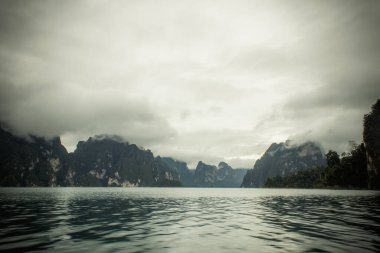 Dağlar, ağaçlar, sis ve önünde bir göl olan manzara, yağmurlu bir günün ardından akşam. Kötü ışıklandırma koşulları