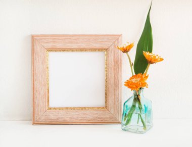 Pastel pembe kare fotoğraf çerçevesi ve turuncu çiçek (kasımpatı) arkaplan olarak beyaz bir duvar ile ahşap bir rafta yeşil yapraklar vazo içinde