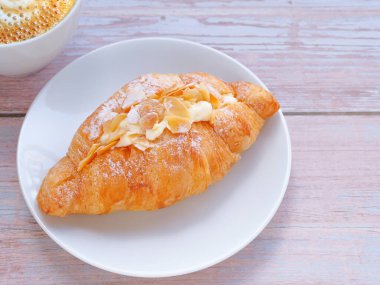 Croissant with cream and almonds on white circle plate and coffee cup over wooden table. Close up, space for text