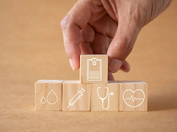 Concept annual health check-up. Hand touch wooden cubes with healthcare medical icons report, blood, syringe, stethoscope, and heartbeat on brown background. health insurance and business background