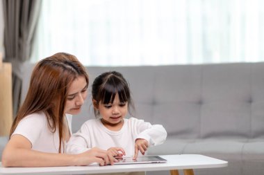 Güzel Asyalı anne ve kızı kanepede oturuyor ve evde birlikte vakit geçirirken bir tablette oyun oynuyorlar.