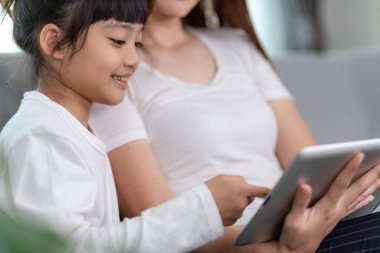 Güzel Asyalı anne ve kızı kanepede oturuyor ve evde birlikte vakit geçirirken bir tablette oyun oynuyorlar.
