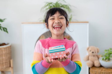 Küçük Asyalı kız gülümsüyor ve oturma odasında kırmızı hediye kutusunu tutuyor. Noel 'de ve yeni yılda hediye kutusunu tutan çocuk..