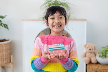 Küçük Asyalı kız gülümsüyor ve oturma odasında kırmızı hediye kutusunu tutuyor. Noel 'de ve yeni yılda hediye kutusunu tutan çocuk..