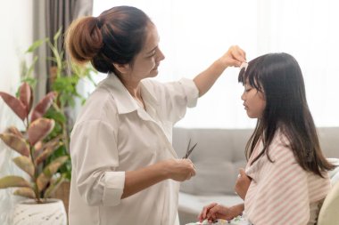 Asyalı anne evde, evinde, Covid-19 Coronavirus 'tan korunurken kızına saç kesiyor. Kişisel karantina ve sosyal uzaklık kavramı.