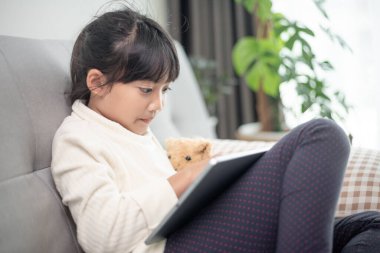 İnternette tablet kullanan küçük bir kız kanepede oturan ya da arkadaşıyla internetten konuşan bir çocuk, sabah oturma odasında dinlenen çocuk, yeni teknoloji konsepti olan çocuklar.