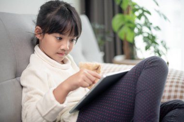 İnternette tablet kullanan küçük bir kız kanepede oturan ya da arkadaşıyla internetten konuşan bir çocuk, sabah oturma odasında dinlenen çocuk, yeni teknoloji konsepti olan çocuklar.