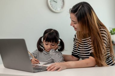 Asyalı küçük kız evde annesiyle online ders alıyor. Anaokulu çocuğu dizüstü bilgisayar kullanır ev ödevi yapar, okul öğretmeninden dijital uzaktan internete geçer ve annesinden destek alır..