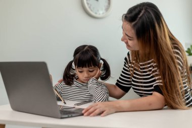 Asyalı küçük kız evde annesiyle online ders alıyor. Anaokulu çocuğu dizüstü bilgisayar kullanır ev ödevi yapar, okul öğretmeninden dijital uzaktan internete geçer ve annesinden destek alır..