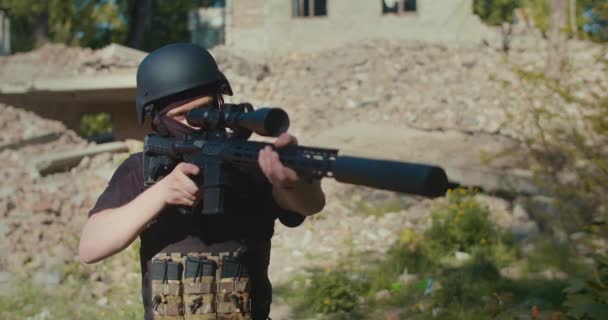 A soldier against the background of a destroyed house, picks up a rifle, aims and makes a shot. — Vídeo de stock