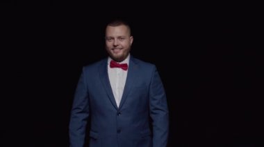 Mavi smokinli, kırmızı kravatlı zarif bir adam gülümser ve kollarını siyah bir arka planda açar..