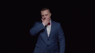 Takım elbiseli bir adam sahnede duruyor ve kameraya bakıyor. Sakalını elleriyle okşuyor..