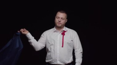 Beyaz tişörtlü, sakallı, seksi bir adam omzunun üzerinden bir ceket atıyor ve siyah arka plana gülümsüyor..