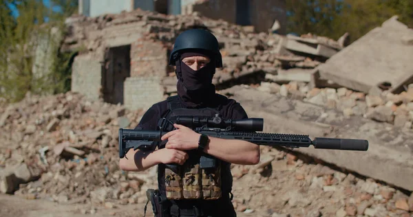 Bir asker yıkılmış bir evin yanında duruyor, yan tarafa bakıyor ve tüfeğini alıyor..