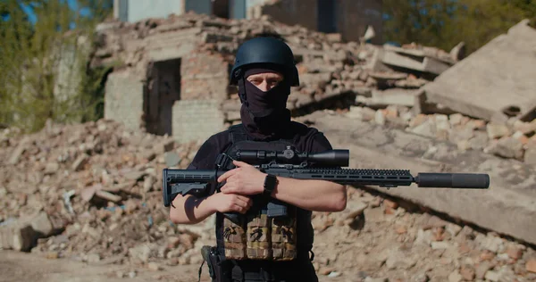 Bir asker yıkılmış bir evin yanında duruyor, yan tarafa bakıyor ve tüfeğini alıyor. Orta boy..