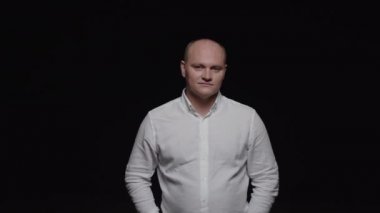 Beyaz tişörtlü zarif kel bir adam parmaklarını vuruyor ve siyah arka planda kameraya poz veriyor..