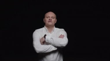 Beyaz gömlekli kel bir adam kollarını kavuşturur ve siyah arka planda kameraya poz verir..