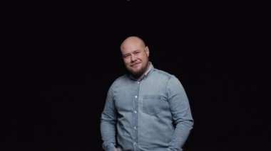 Sakallı kel bir adam kameranın önünde poz veriyor ve siyah bir arka plana gülümsüyor..