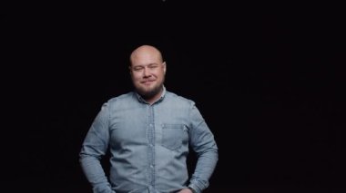 Kot gömlekli kel bir adam gülümsüyor ve siyah arka planda kameraya poz veriyor..