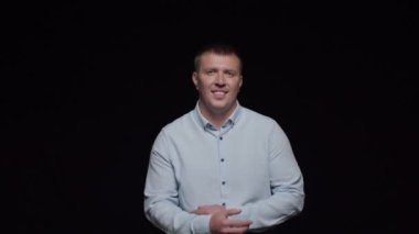 Mavi gömlekli genç bir adam siyah arka planda duruyor ellerini katlıyor ve tatlı tatlı gülümsüyor..