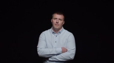 Gömlekli genç bir adam siyah arka planda poz verir, kollarını kavuşturur ve çenesini sıvazlar..