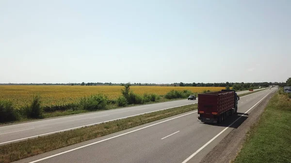 Kargo karavanıyla birlikte yolda giderken ve malları taşırken çekilen hava görüntüsü. Kargo kamyonu üzerinde uçuyor. Otoyol boyunca ilerliyor. Kırsal alanların yakınından geçiyor. Manzaralı doğa sahnesi. Yan görünüm
