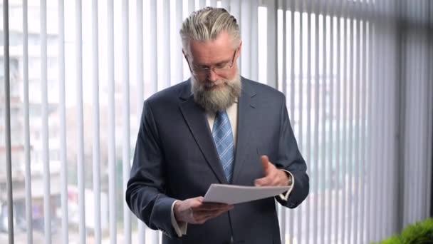Een volwassen, grijsharige baas bestudeert de rapporten en knikt met zijn hoofd in goedkeuring — Stockvideo