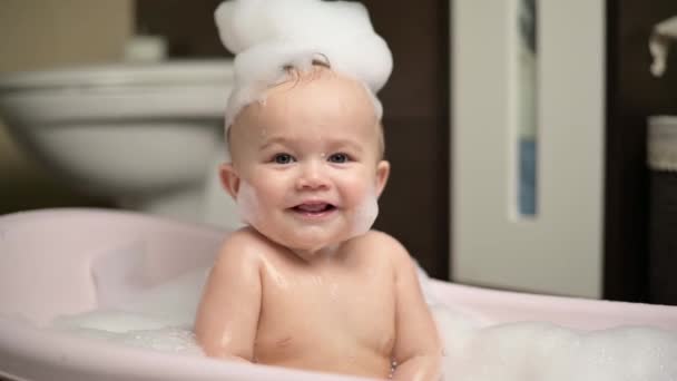 Little boy sits in a bathtub with foam and smiles charmingly — Stockvideo
