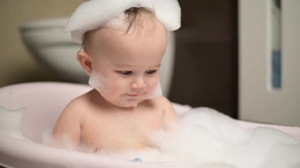 A little boy sits in a bath with foam and looks upward cutely, turning his head — 비디오
