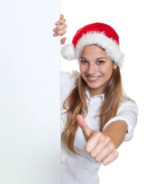 有魅力的女人，戴着圣诞帽子展示招牌后面的拇指 — 图库照片