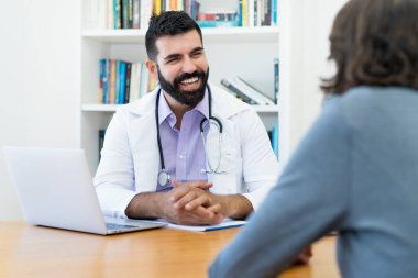 Hippi sakallı yakışıklı bir doktor hastanenin ofisinde sağlıklı bir hastayla konuşuyor.