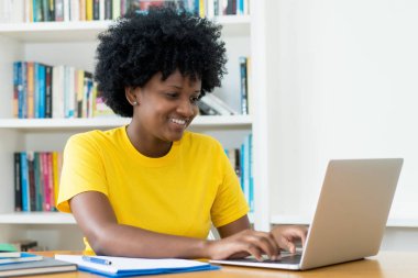 Gülen Afrikalı Amerikalı kız öğrenci evdeki masada bilgisayardan öğreniyor.