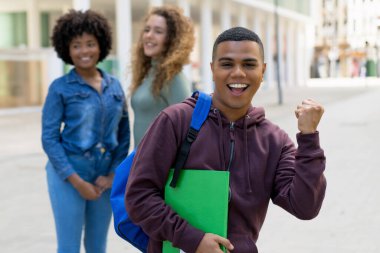Successful cheering latin american male student with backpack and group of international students outdoor in summer in city