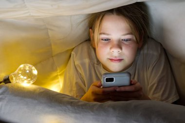 Geceleri battaniyenin altında cep telefonu olan bir kızın portresi. Gadget gecesi. Sosyal medya bağımlılığı. Çevrimiçi iletişim.