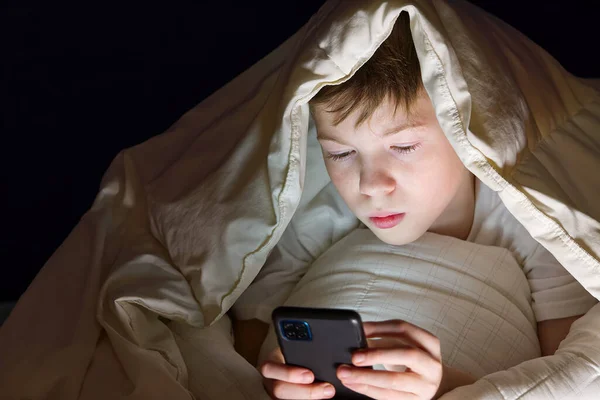 Menino debaixo do cobertor à noite em sua cama se comunica na Internet. Vício em gadgets infantis e insônia. vício em redes sociais — Fotografia de Stock