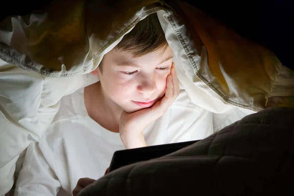 Ensino à distância. Educação online. Menino feliz na cama sob cobertor com tablet no escuro. A cara de crianças ilumina-se pelo monitor brilhante. Quarentena. vício em redes sociais — Fotografia de Stock