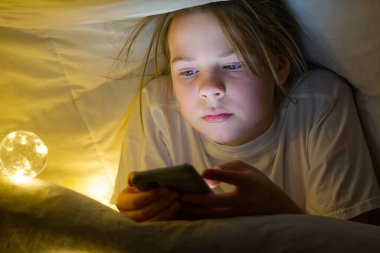 Geceleri battaniyenin altında cep telefonu olan bir kızın portresi. Gadget gecesi. Sosyal medya bağımlılığı. Siber zorbalık